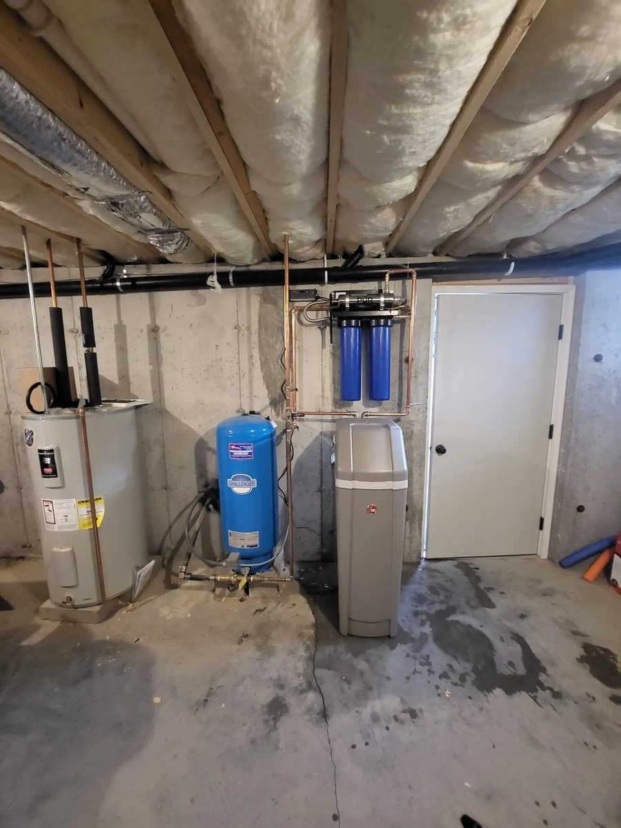 Electric water heater beside a blue well pressure tank and water filter housings in an unfinished basement