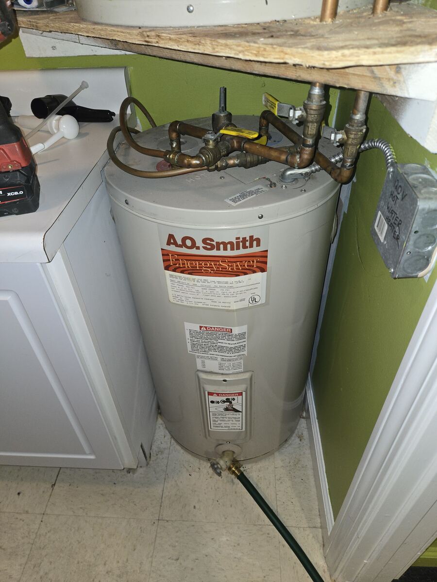 Small electric water heater tucked under a wooden shelf beside a washing machine