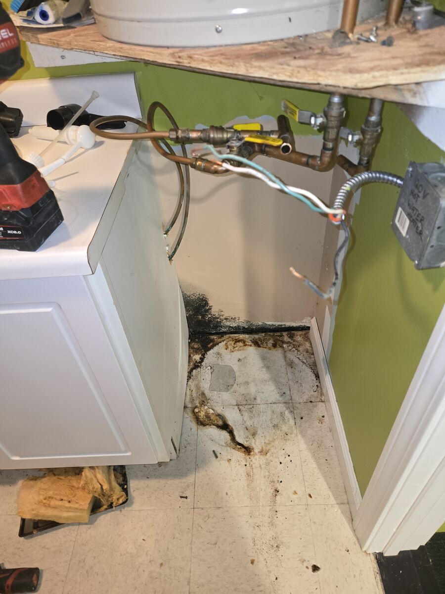 Empty corner after a water heater was removed, showing a stained and damaged floor